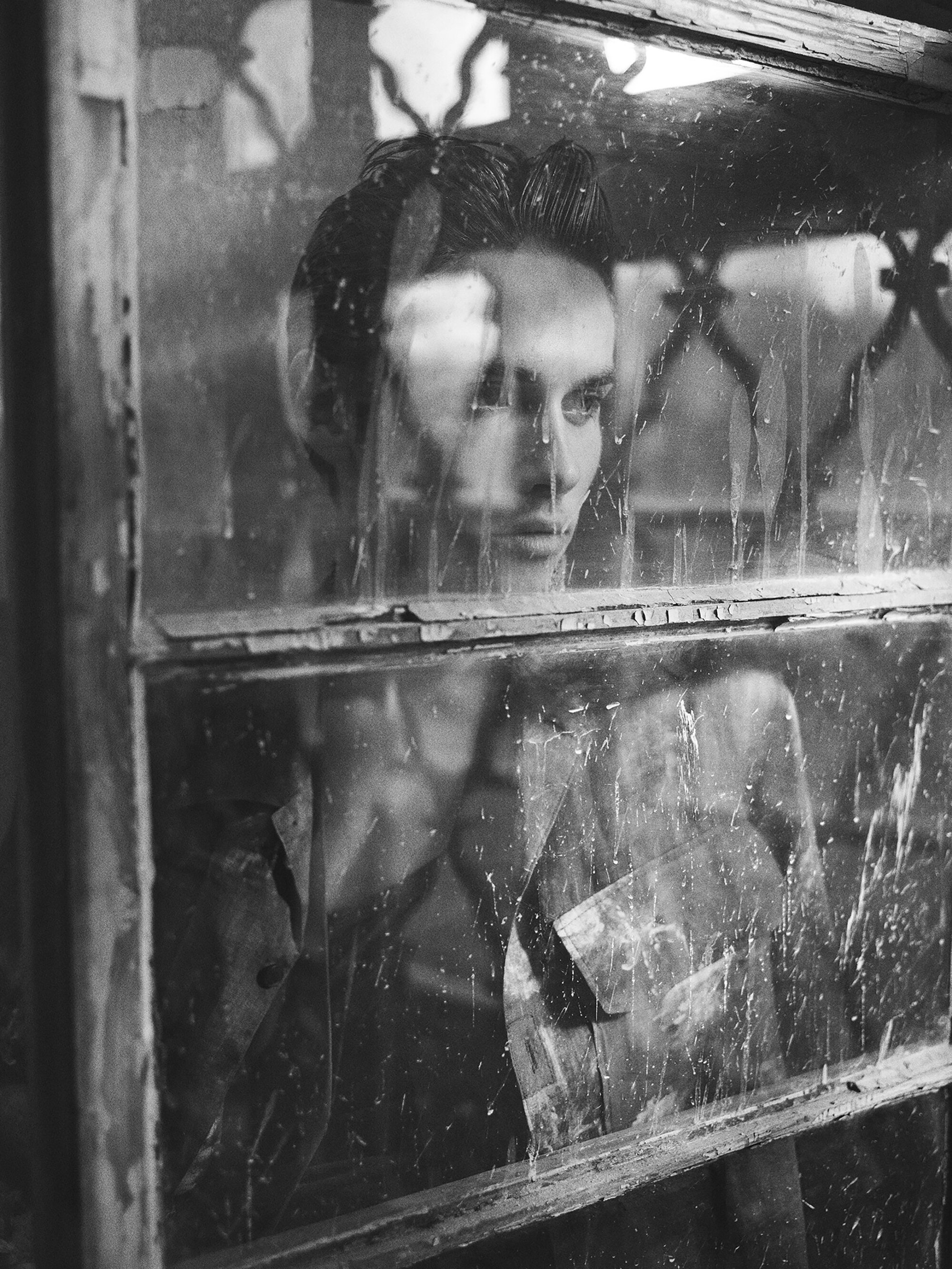 Monochrome portrait of a woman behind a weathered window with damaged glass, conveying a sense of reflection and quiet introspection.