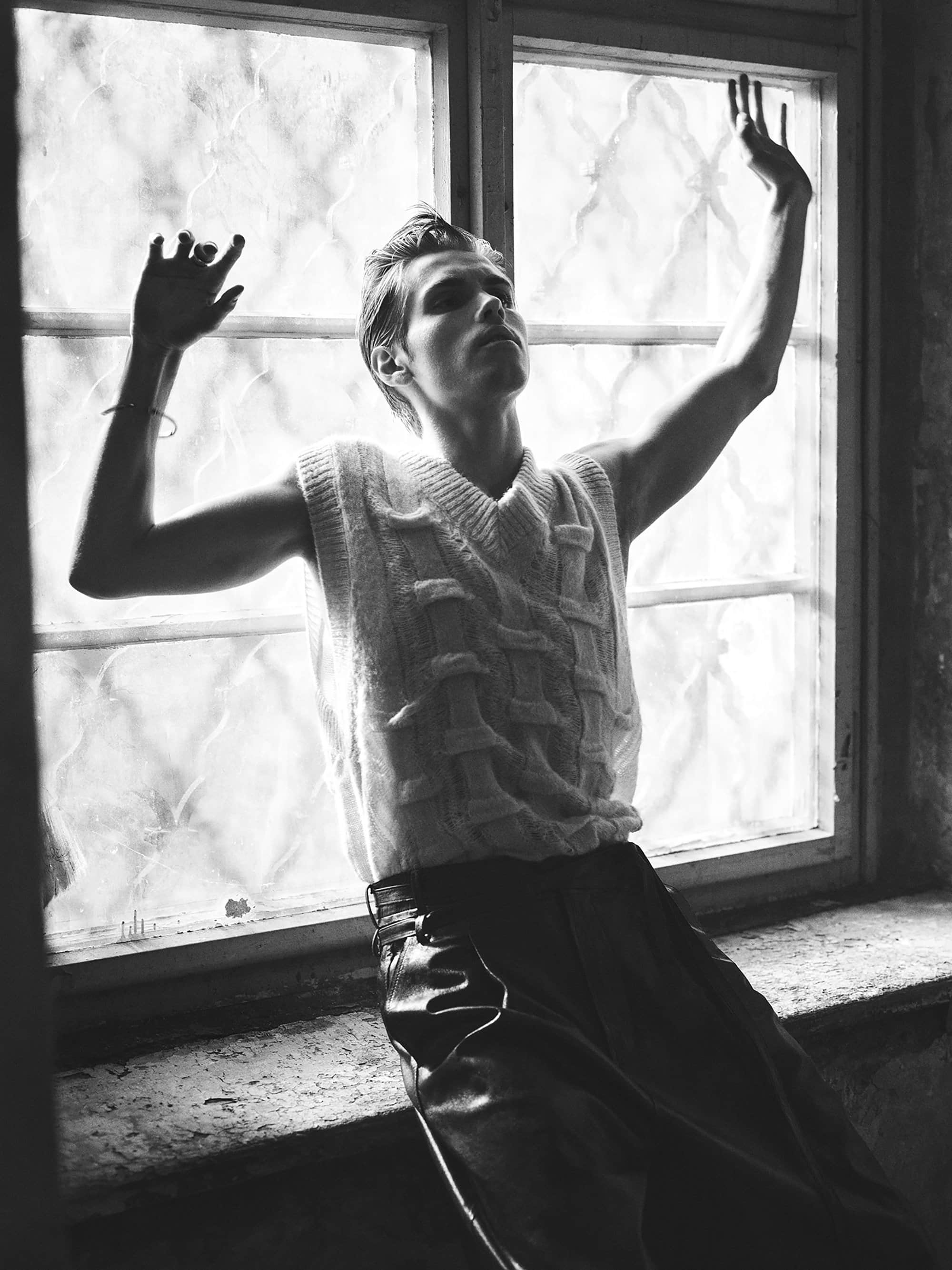 Black and white portrait of a young adult man posing in front of a window. He is wearing a sweater vest and trousers.