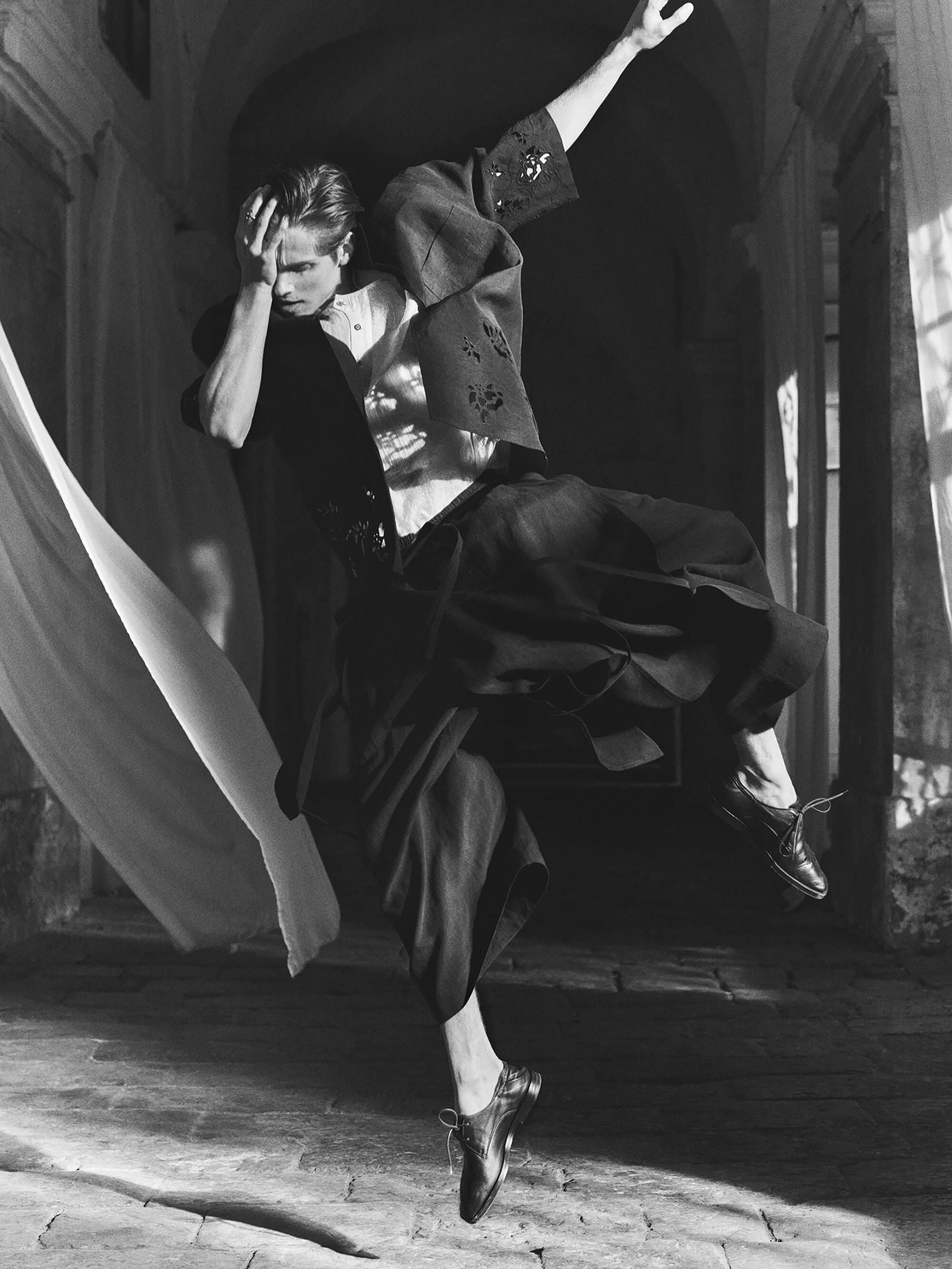 Monochrome fashion shot of a man in mid-air, wearing a floral-patterned shirt and wide pants, with a draped fabric and old stone archway.