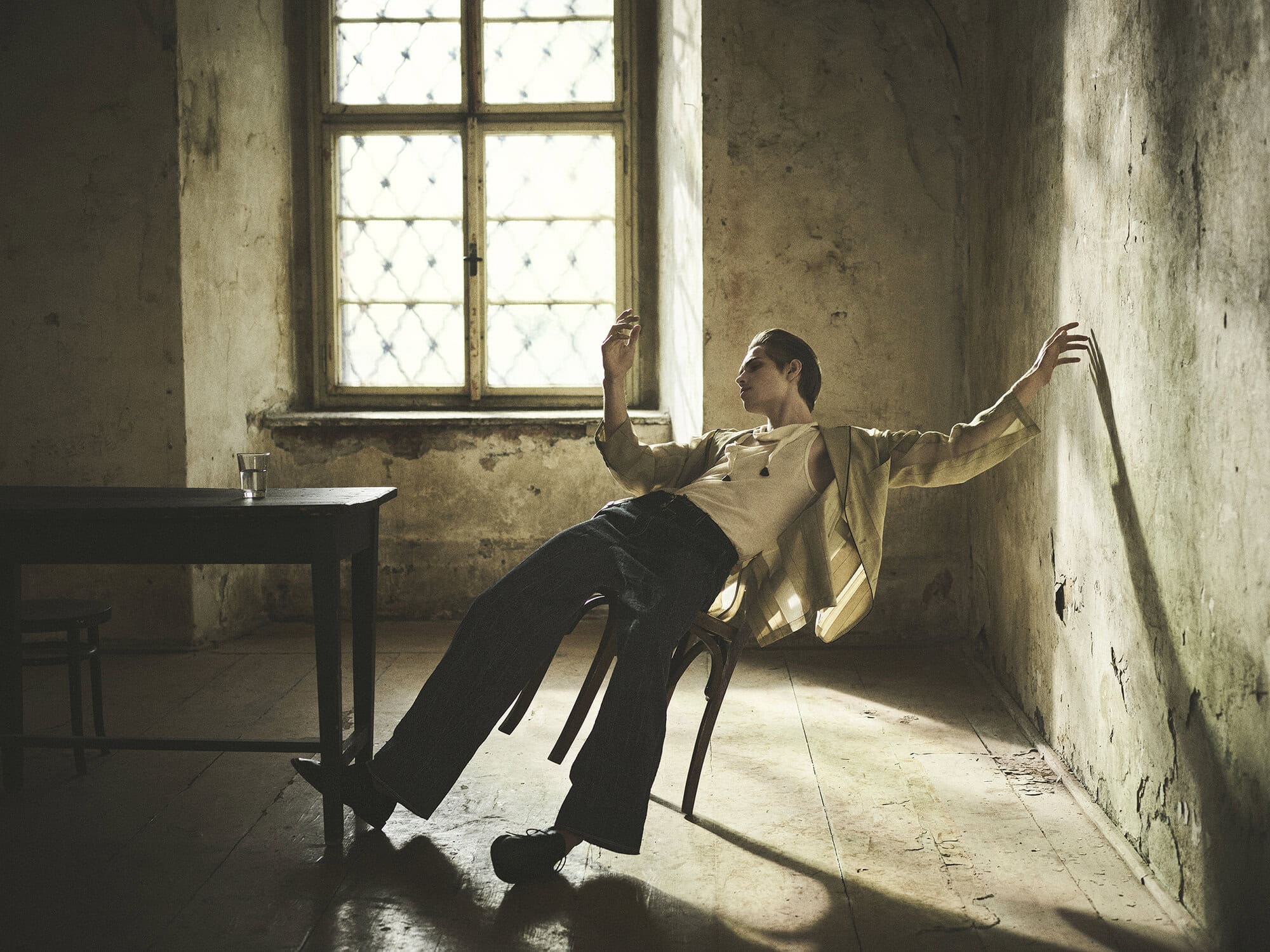 A man leaning back on a wooden chair in a rustic room with a window, wearing a patterned jacket and dark trousers for Zara.
