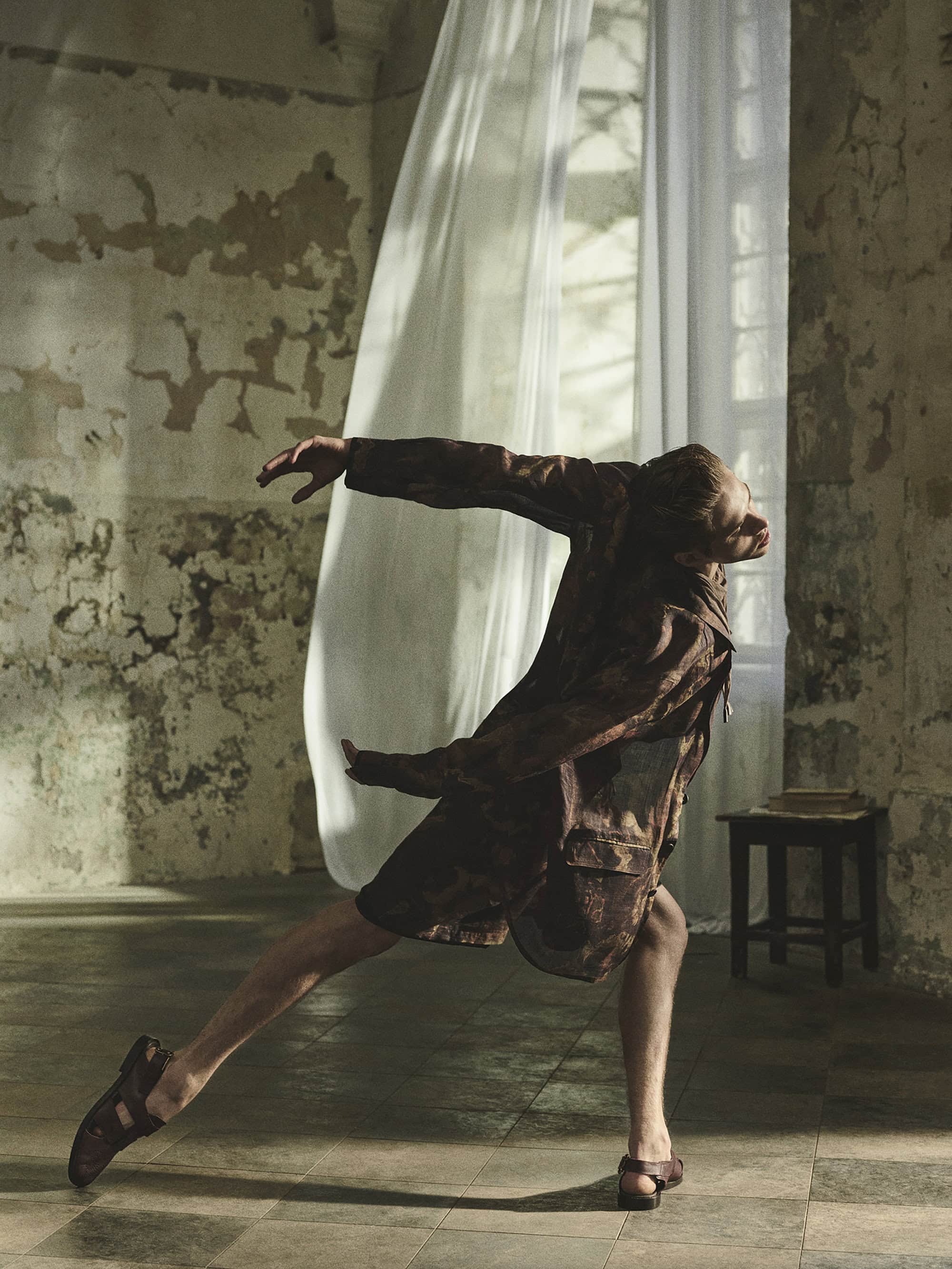 Man dances in rustic interior near a window with white curtains, wearing a patterned brown jacket, shorts, and sandals in elegant pose.