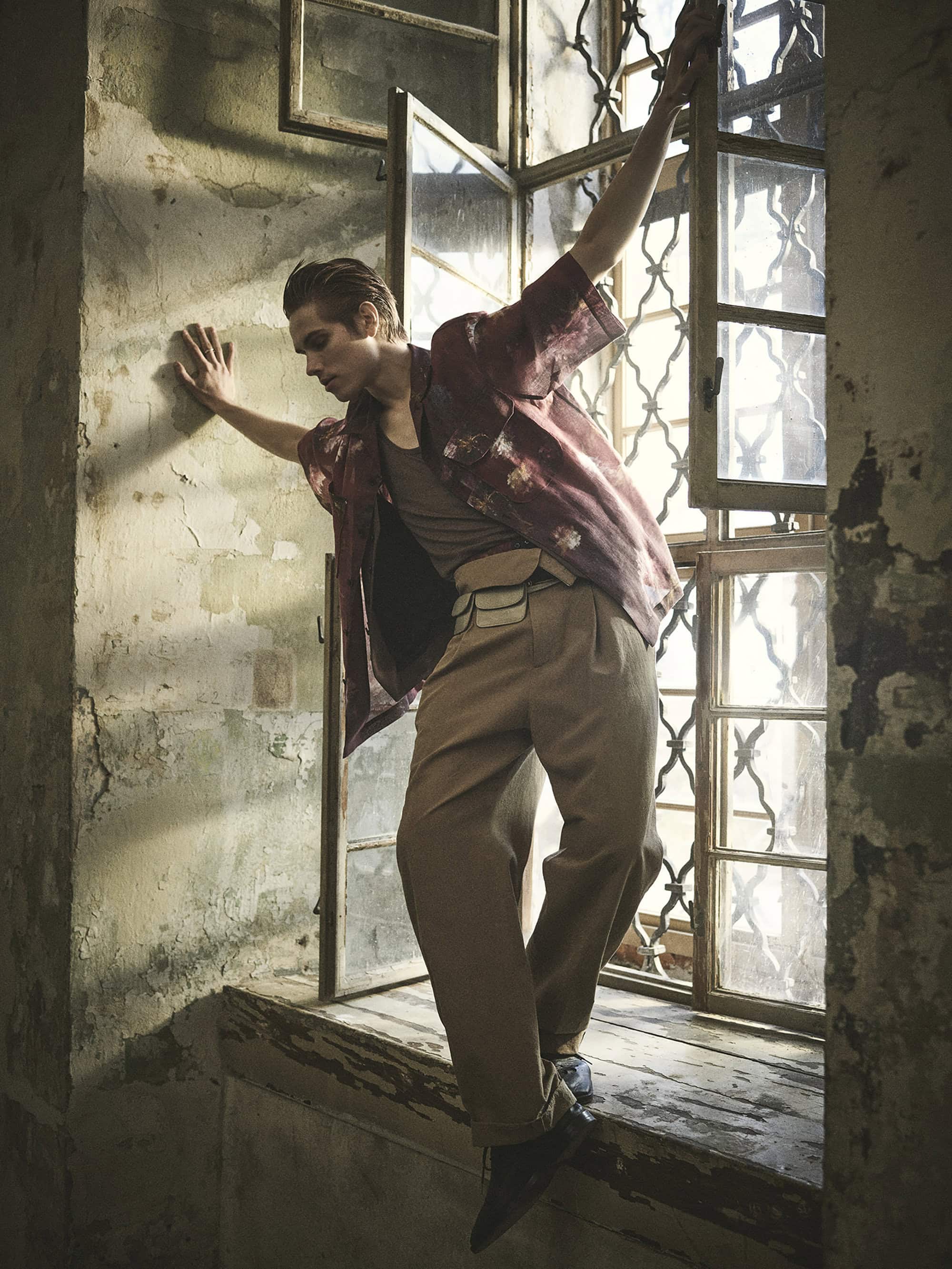 A young adult male in an open window frame wearing a patterned shirt and tan trousers poses in a dimly lit room with weathered walls.