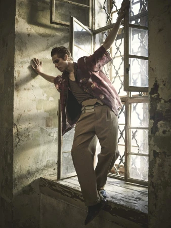 A young adult male in an open window frame wearing a patterned shirt and tan trousers poses in a dimly lit room with weathered walls.