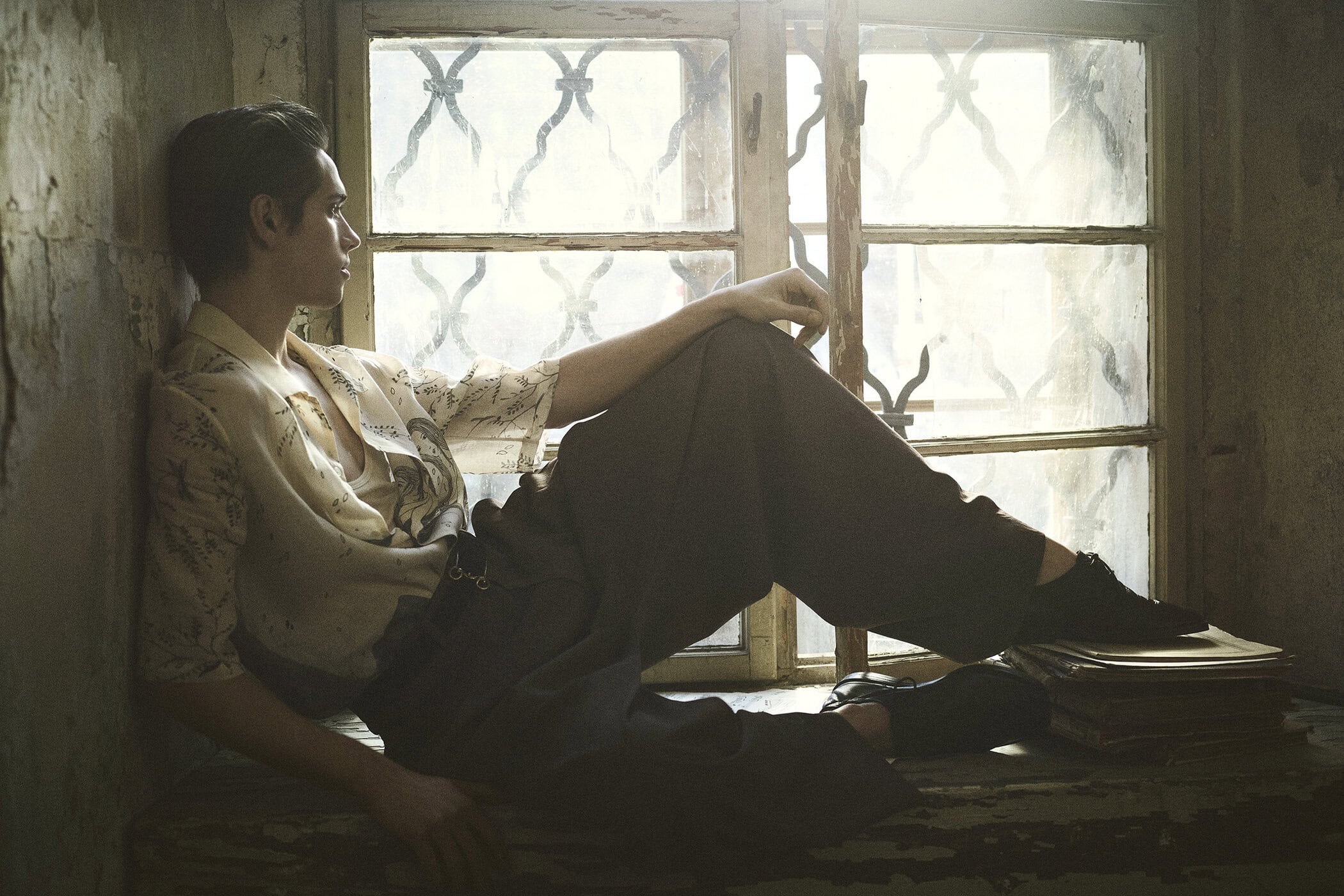 Man in patterned shirt and dark trousers poses seated on windowsill, bathed in soft light from window. Books visible below, neutral tones.