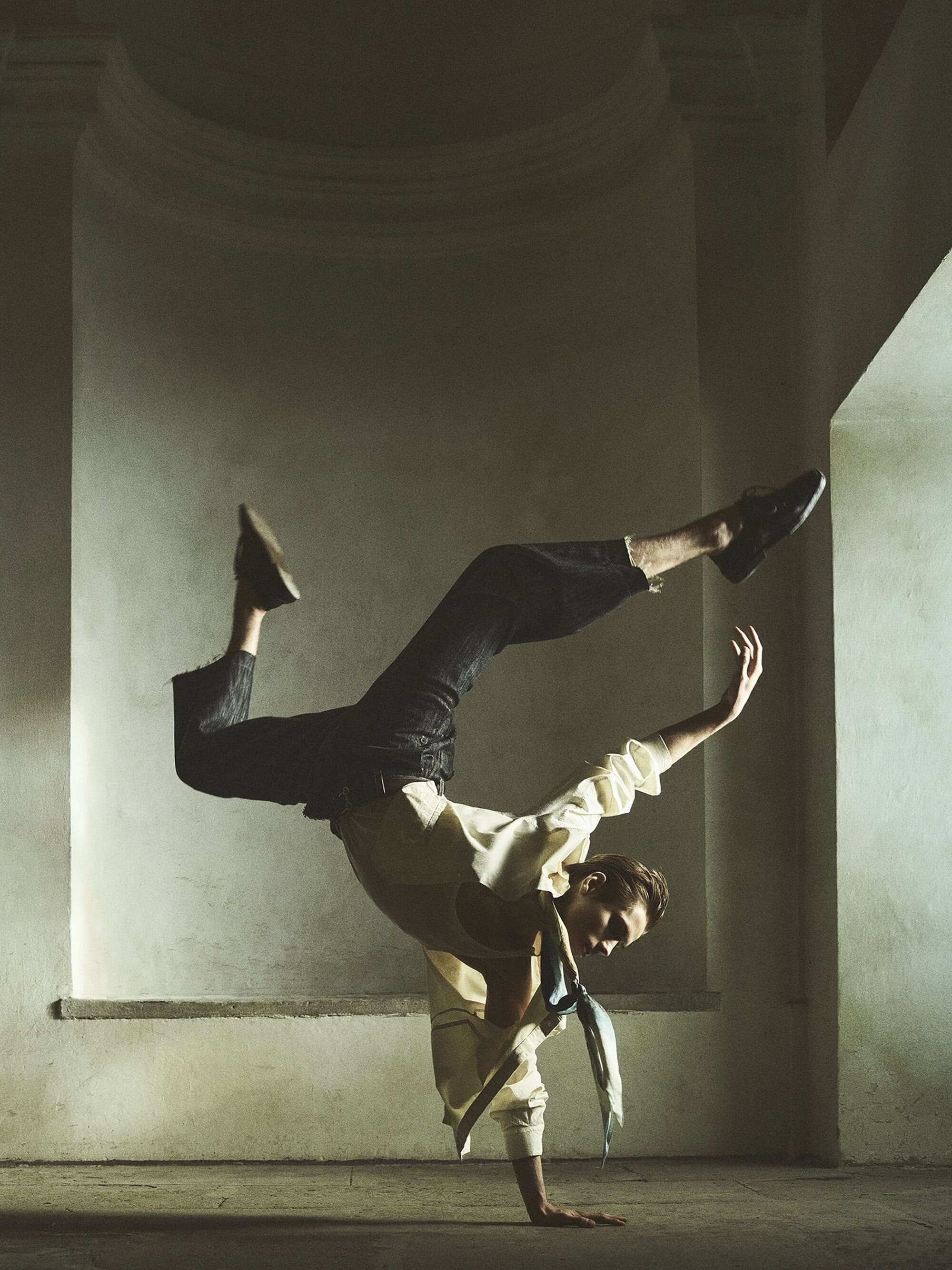 Adult male dancer in a handstand pose wearing a white shirt, dark trousers, and shoes in an indoor architectural space.