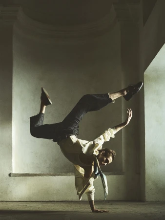 Adult male dancer in a handstand pose wearing a white shirt, dark trousers, and shoes in an indoor architectural space.
