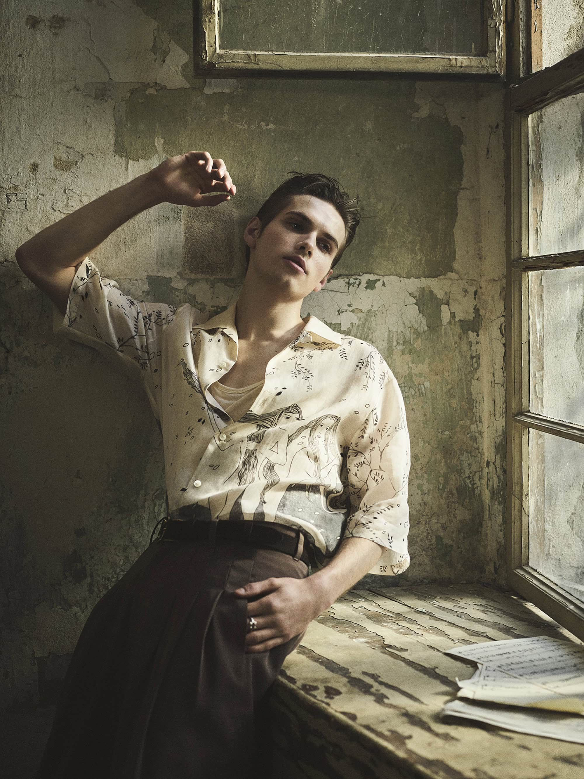Portrait of a young adult man in printed shirt posing indoors near a window, leaning against a textured wall with natural lighting.