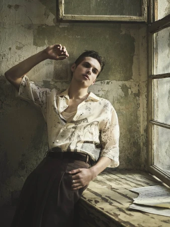 Portrait of a young adult man in printed shirt posing indoors near a window, leaning against a textured wall with natural lighting.