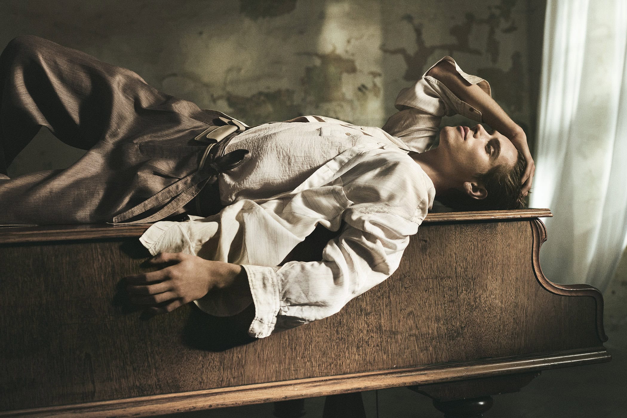 Portrait of an adult male wearing loose clothing, lying on a vintage wood piano indoors, illuminated by soft light, against a textured wall.