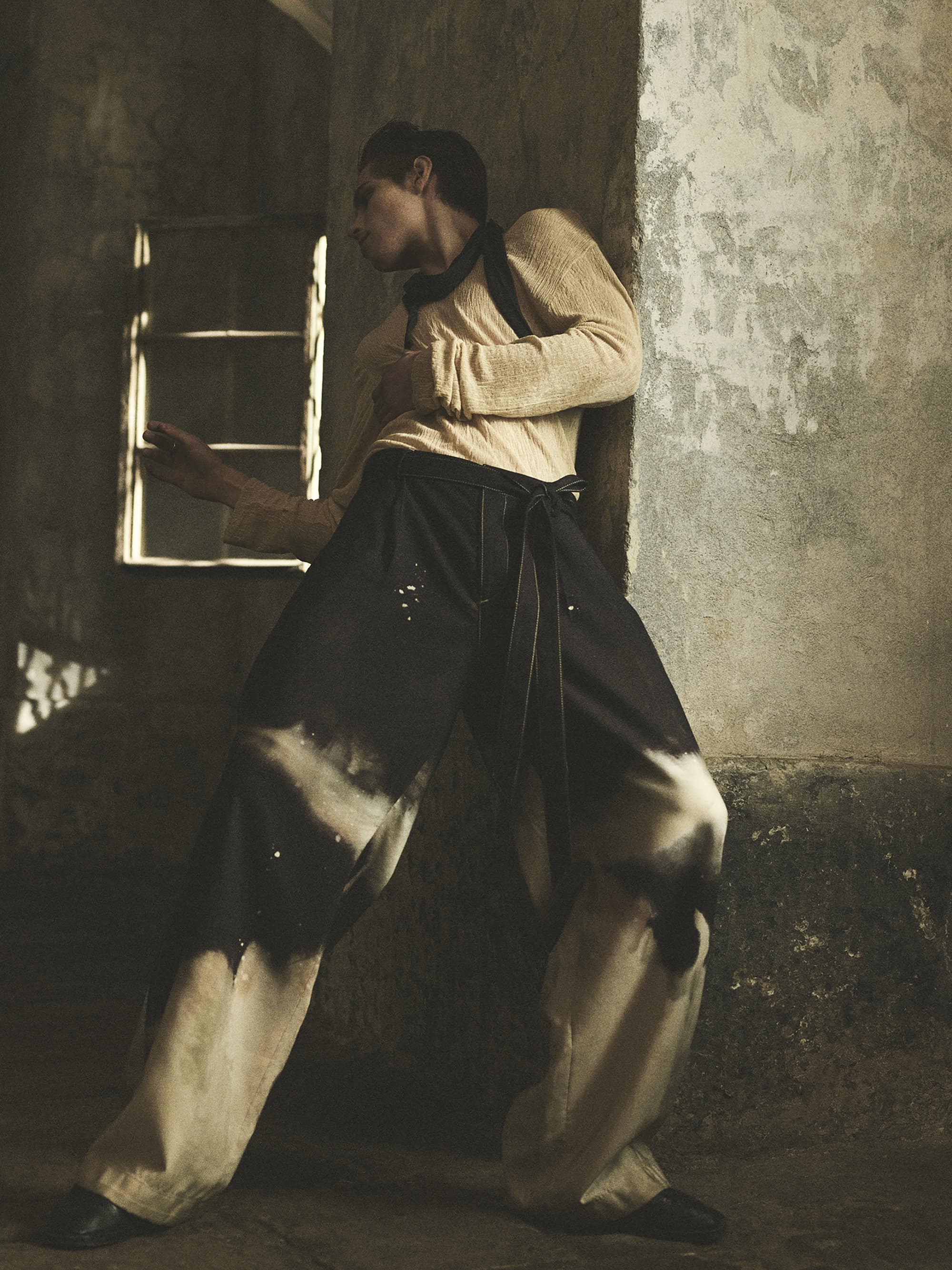 A slender adult wearing wide, tie-waist pants poses indoors next to a textured wall and a shadowed window, illuminated by soft, diffused lighting.