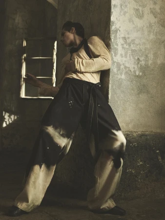 A slender adult wearing wide, tie-waist pants poses indoors next to a textured wall and a shadowed window, illuminated by soft, diffused lighting.