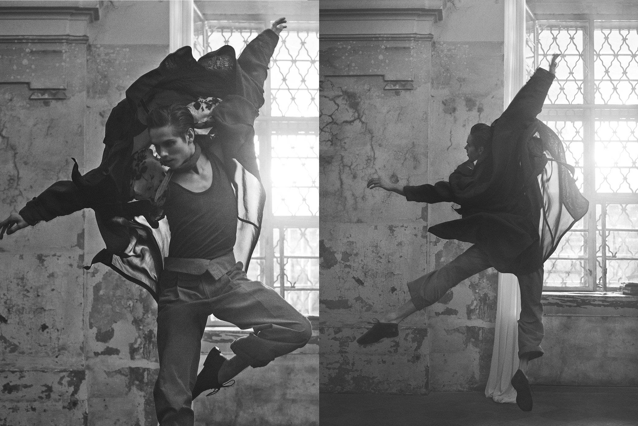 Two adult male dancers, one in a tank top and the other in a coat, are frozen mid-air in a derelict, grayscale interior with light from windows.