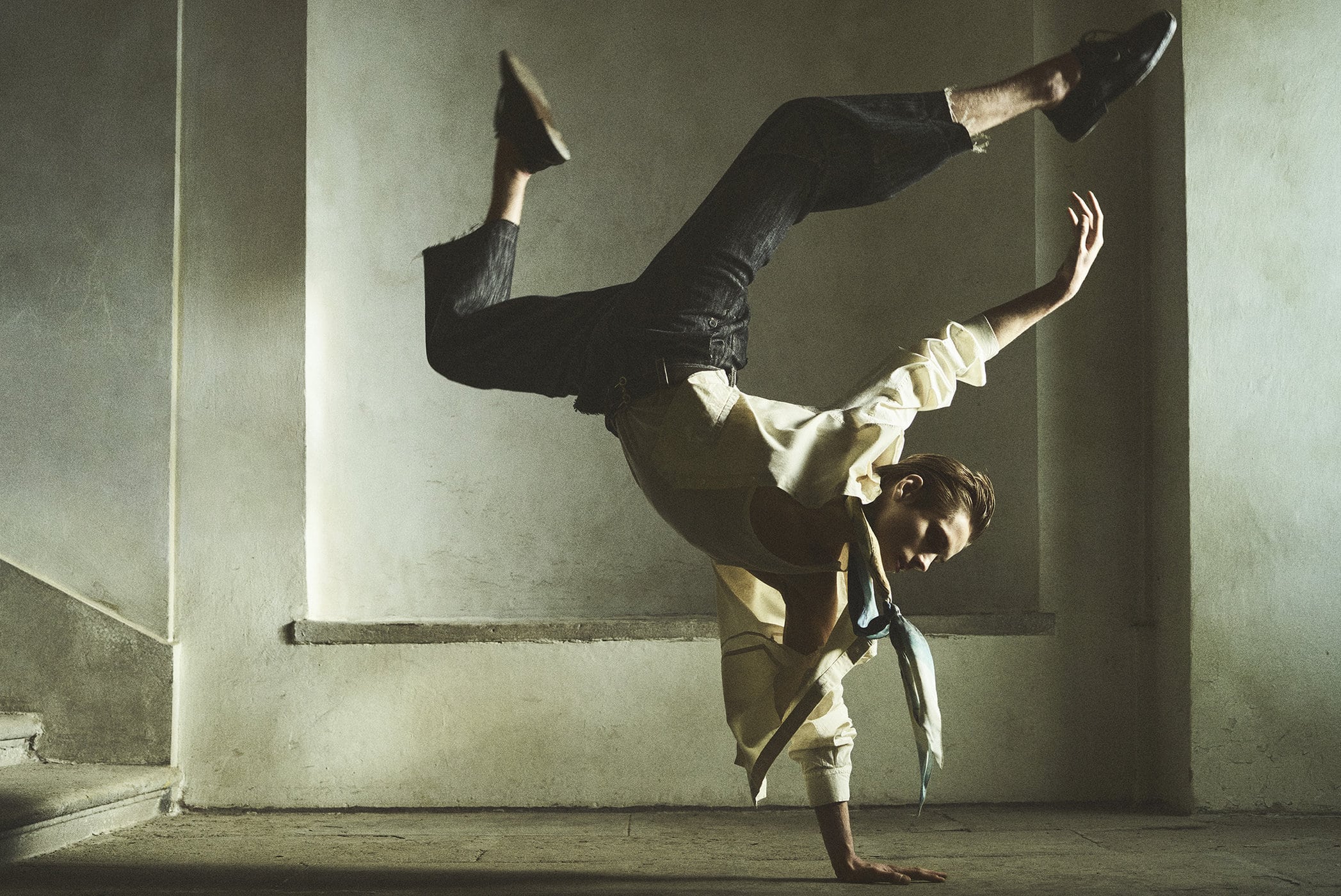 A person is doing a handstand in an indoor setting with concrete walls, wearing a shirt, pants, and shoes, lit by natural light.