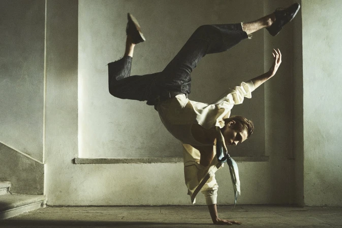 A person is doing a handstand in an indoor setting with concrete walls, wearing a shirt, pants, and shoes, lit by natural light.
