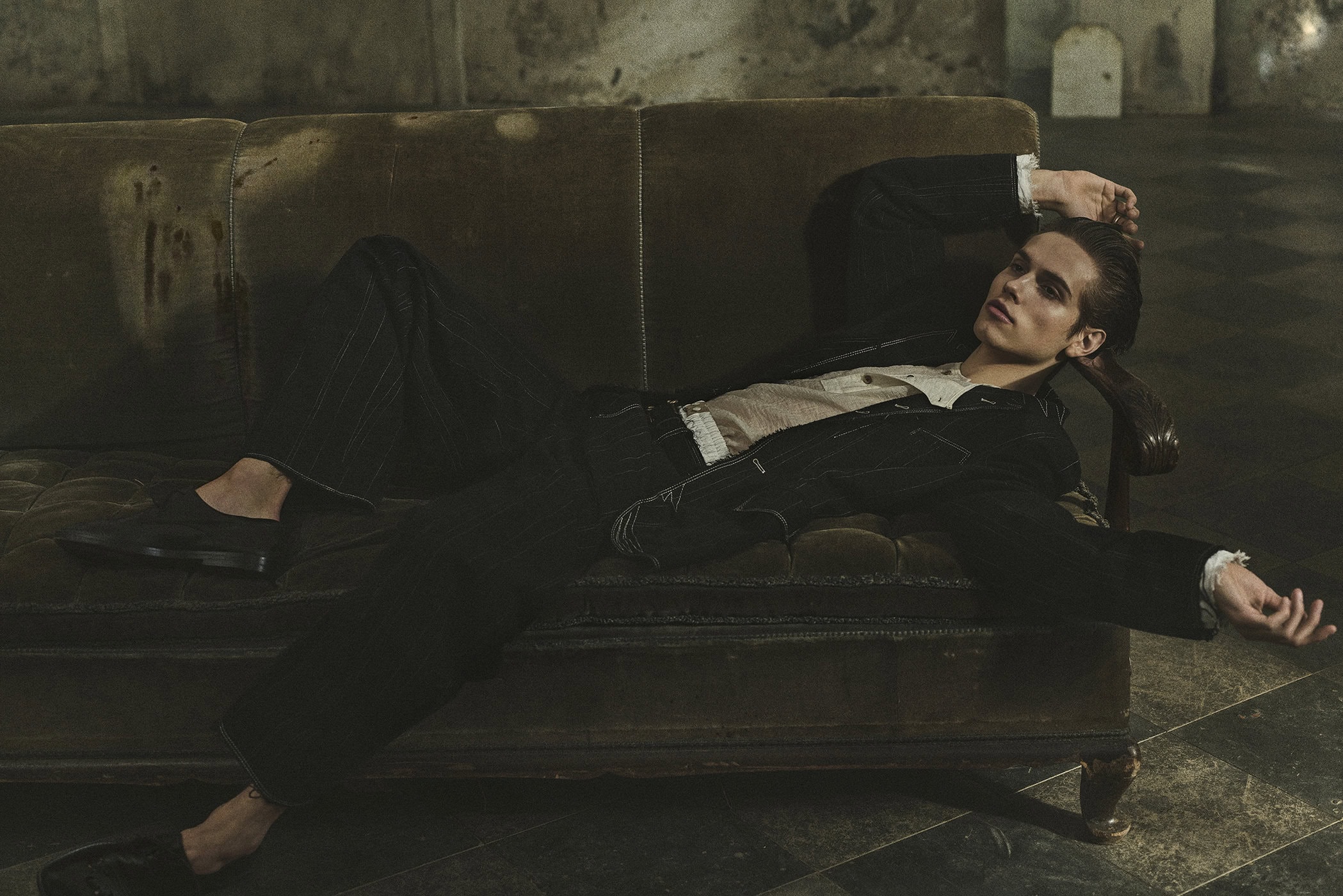 A young adult male model poses on a vintage sofa in a dimly lit room with dark walls and a patterned floor, wearing a jacket and pants.