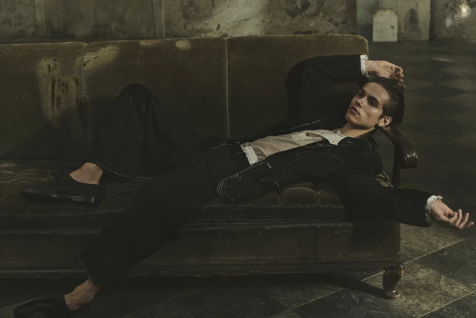 A young adult male model poses on a vintage sofa in a dimly lit room with dark walls and a patterned floor, wearing a jacket and pants.