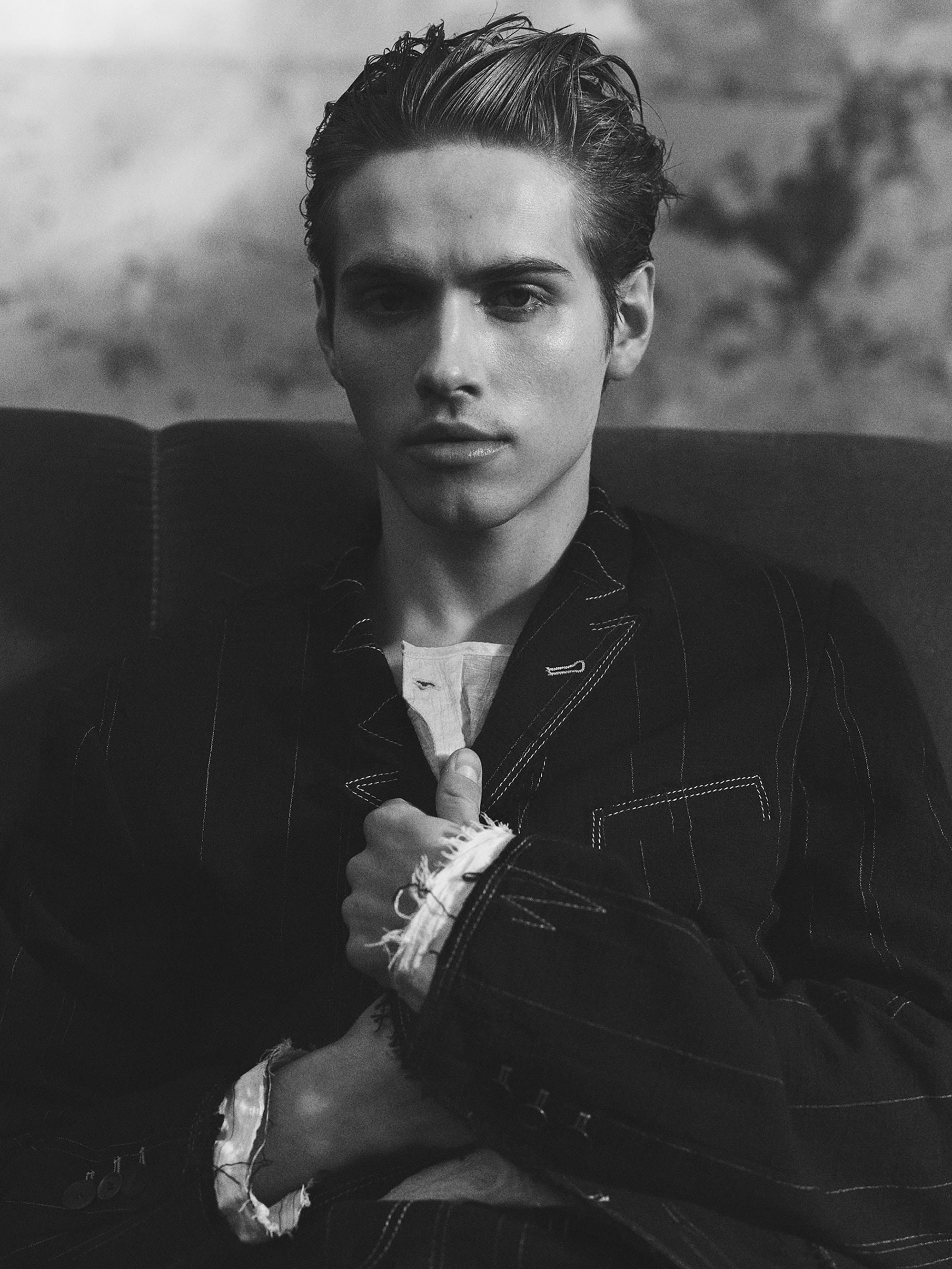 Portrait of a handsome adult male with slicked-back hair, wearing a pinstripe jacket and white shirt, posing against a textured background.