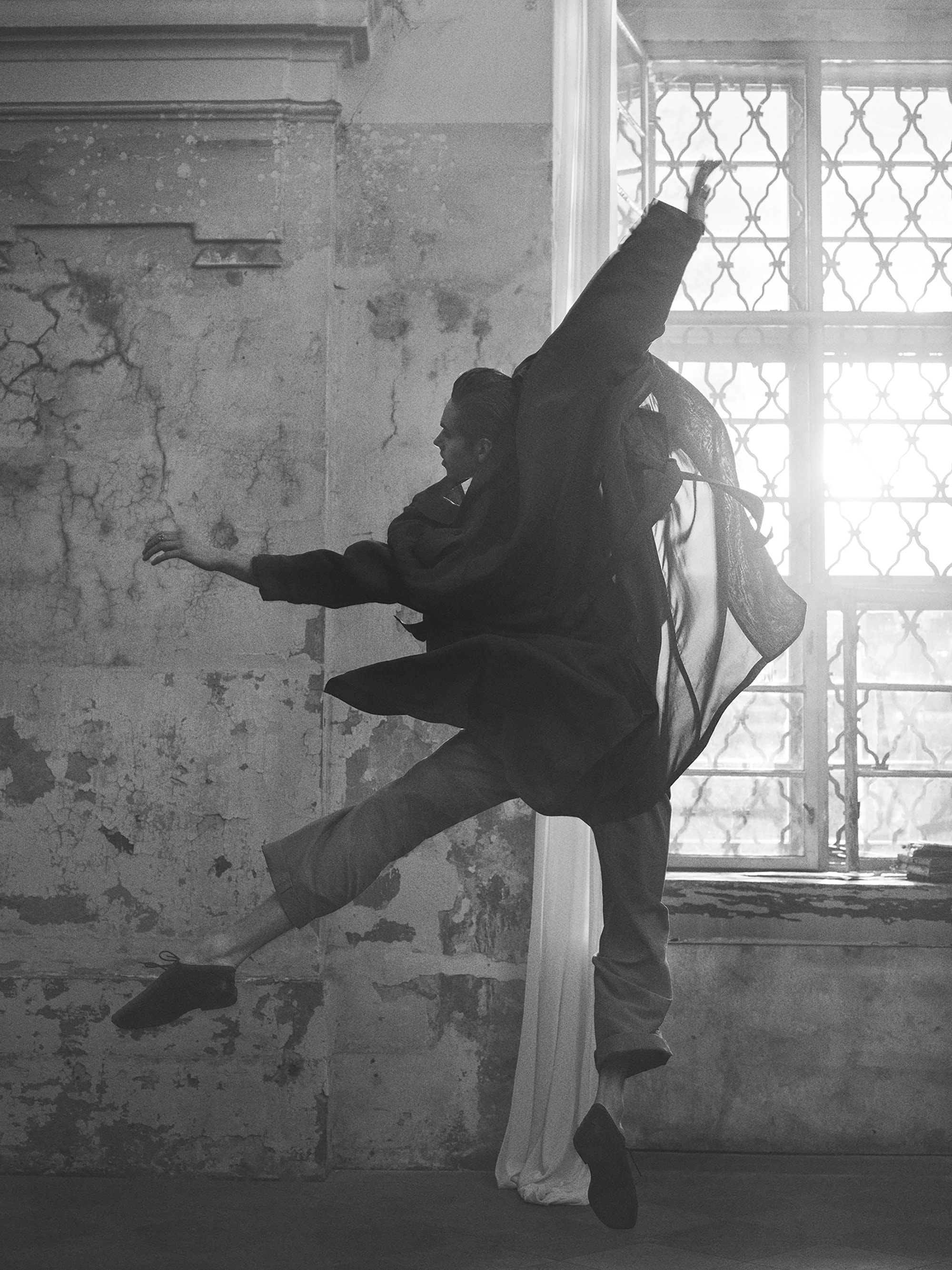 Monochrome image of a person mid-air, wearing a coat, pants, and ballet shoes, in a room with large windows and aged walls.