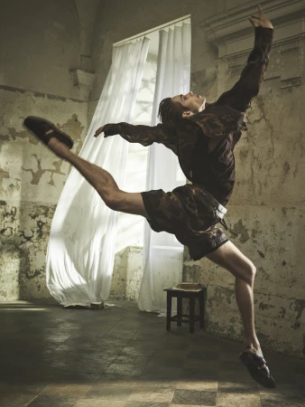 Male ballet dancer leaps in old-world dance studio near bright window light, creating artistic fashion editorial and fitness scene.
