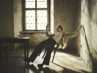 Model leaning backward on a wooden chair in a rustic room with a window, photographed by Annemarieke van Drimmelen for Zara.
