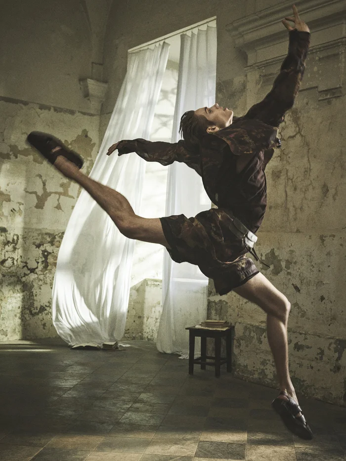 Dancer in dark garments captured mid-air in a high leap inside a room with rustic textured walls and a sheer white curtained window
