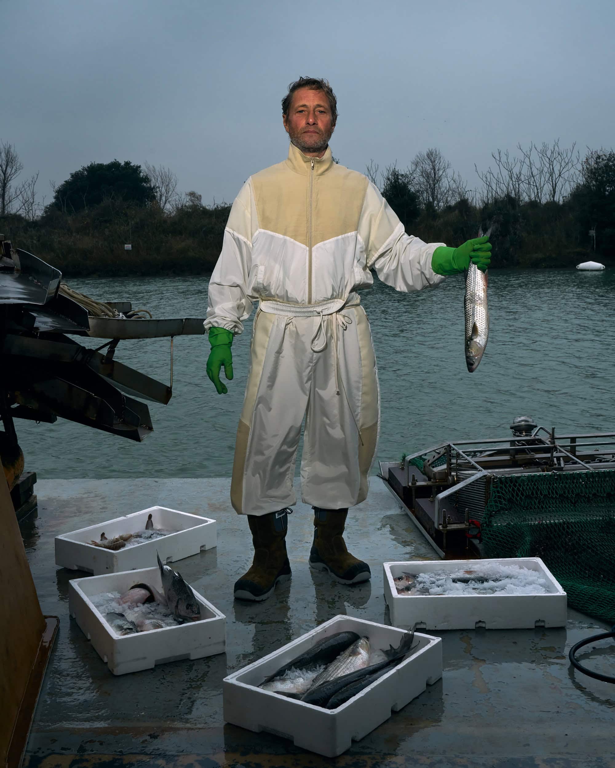 Fisherman displays a freshly caught fish on a boat, surrounded by boxes of fish and river background, captured in a medium outdoor shot.