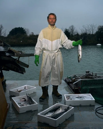 Fisherman displays a freshly caught fish on a boat, surrounded by boxes of fish and river background, captured in a medium outdoor shot.