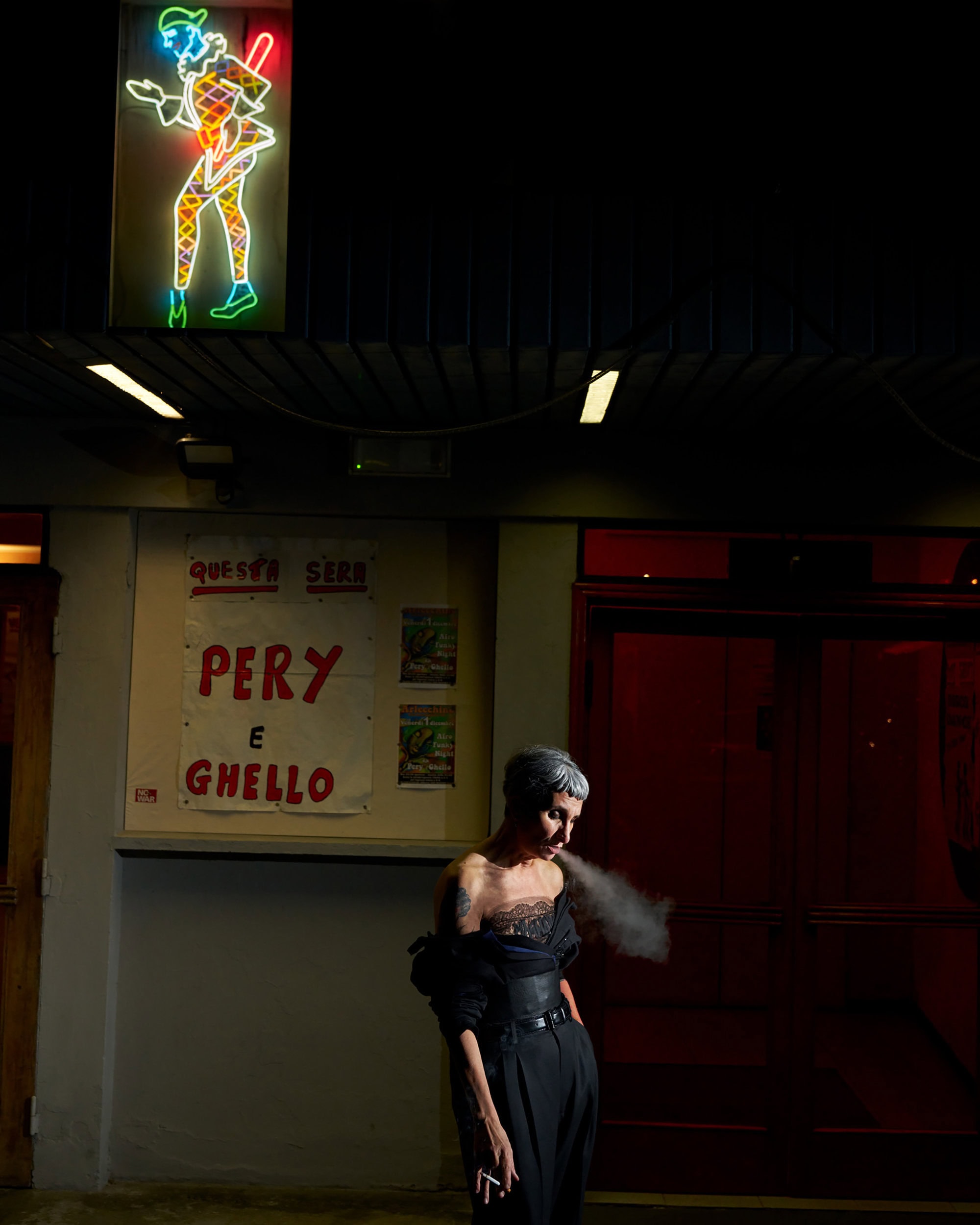 Woman exhales smoke in front of a building with a neon clown sign, captured in an editorial style at night.