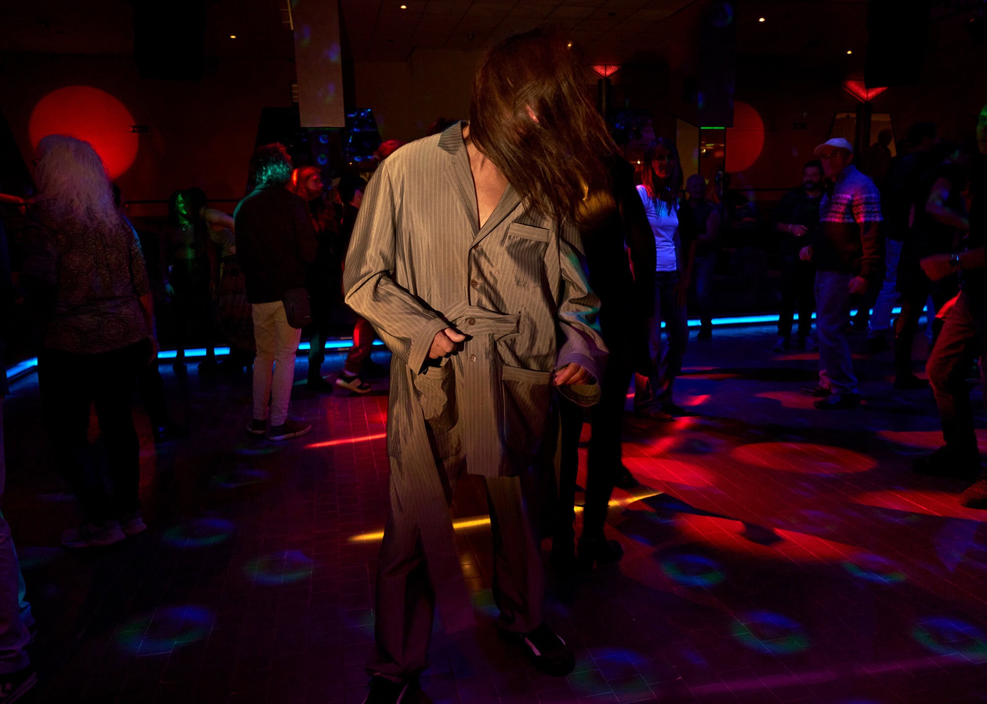 A man in gray pajamas dancing in a dimly lit club with red and blue lights, surrounded by other people on the dance floor.