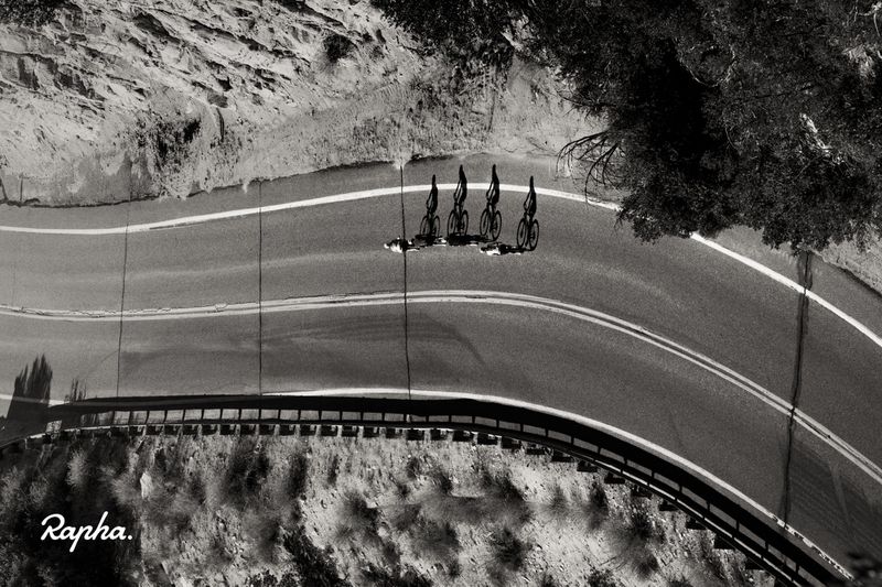 Black and white aerial shot of four cyclists riding down a curving mountain road next to trees and rocky terrain.