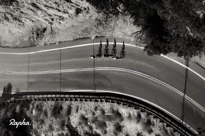Black and white aerial shot of four cyclists riding down a curving mountain road next to trees and rocky terrain.