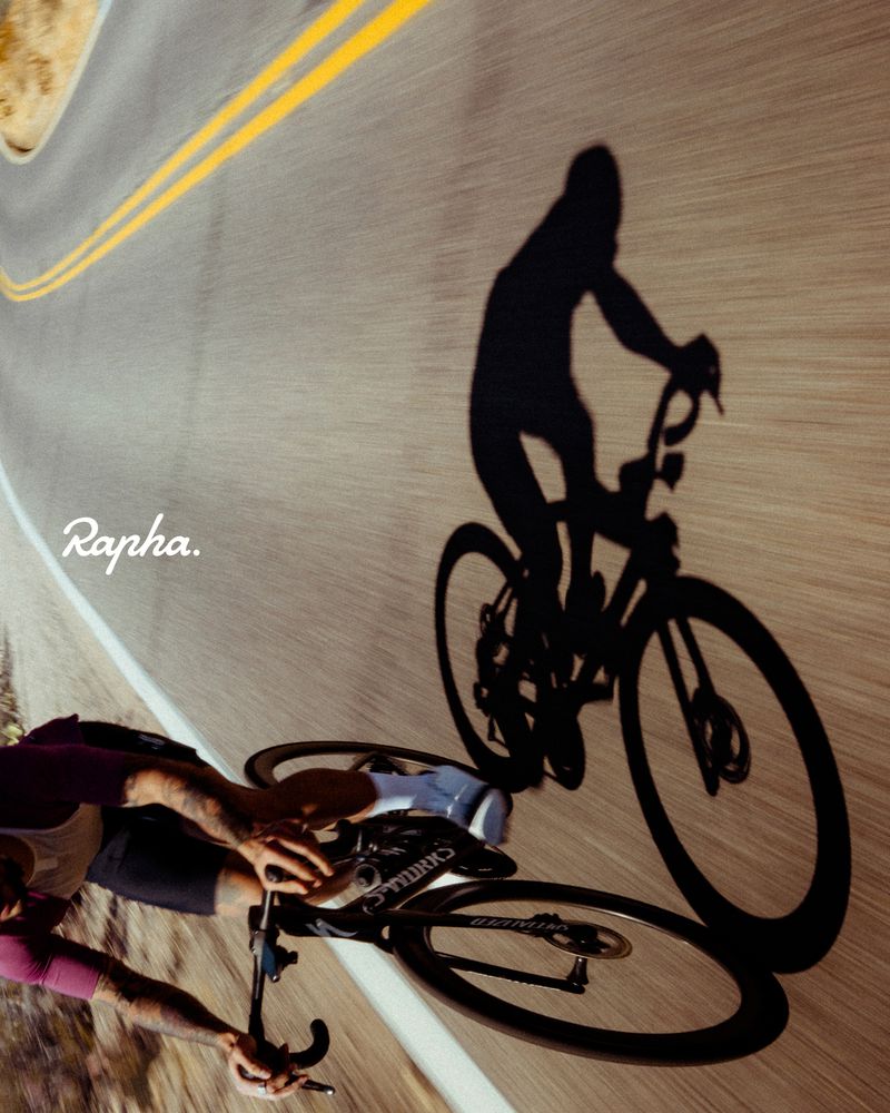 Low-angle shot of a cyclist riding on an asphalt road with double yellow lines, casting a long shadow, next to the Rapha logo.