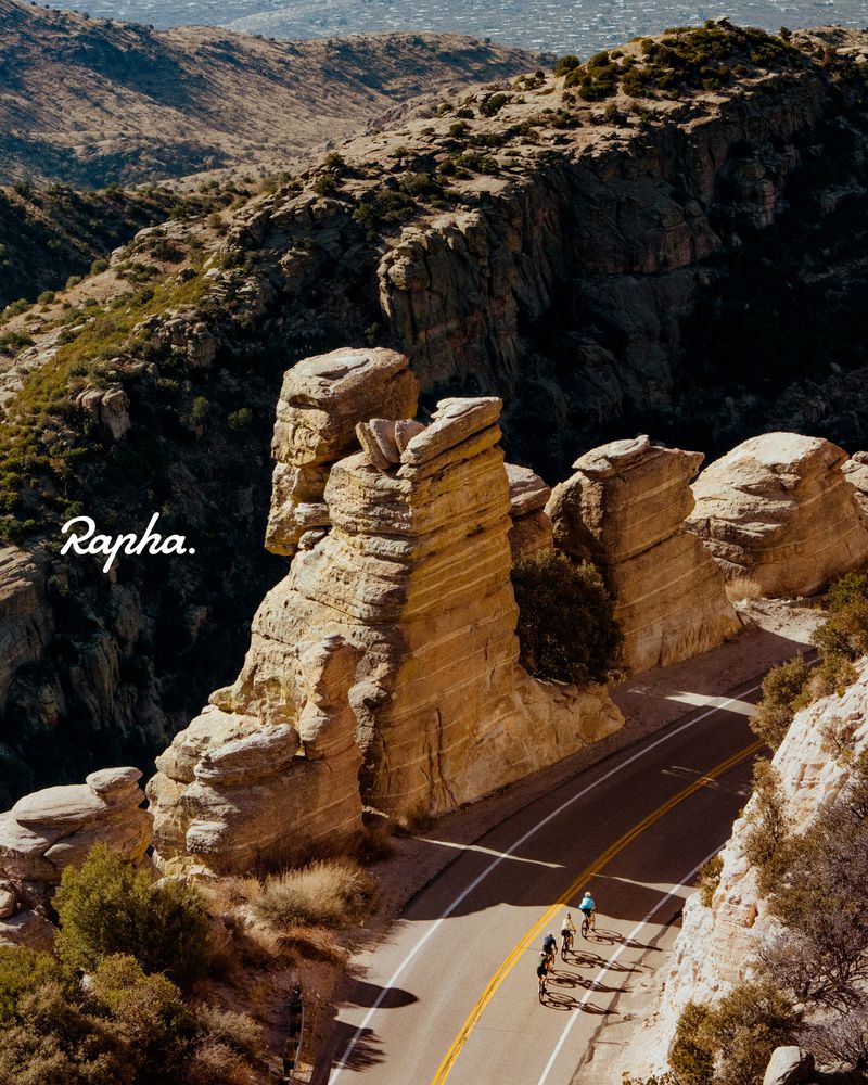 Scenic view of cyclists riding on a mountain road framed by rock formations with the Rapha logo visible, set against a desert backdrop.