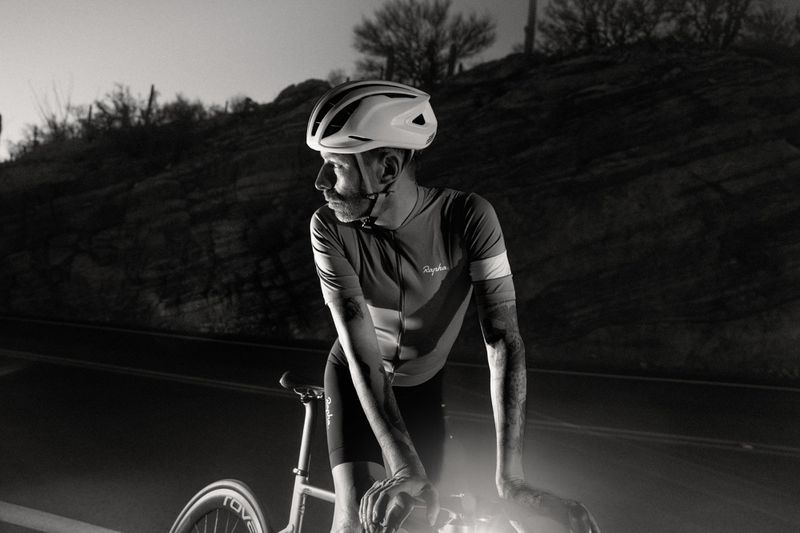 Monochrome shot of a cyclist in a helmet leaning on his bike on a road with dark hills in the background in dim lighting.