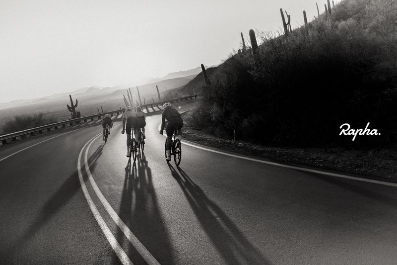 Black and white shot of cyclists riding along a winding desert road, with cacti dotting the hillside and the Rapha logo in the corner.