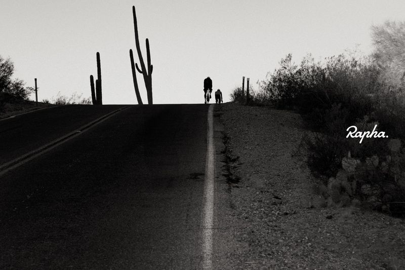 Cyclist and dog silhouetted on a desert road with cacti under a bright sky, creating a stark black and white image.