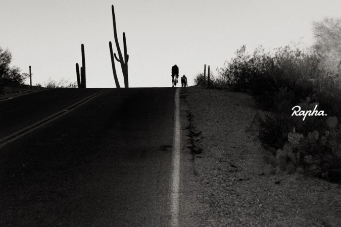 Cyclist and dog silhouetted on a desert road with cacti under a bright sky, creating a stark black and white image.