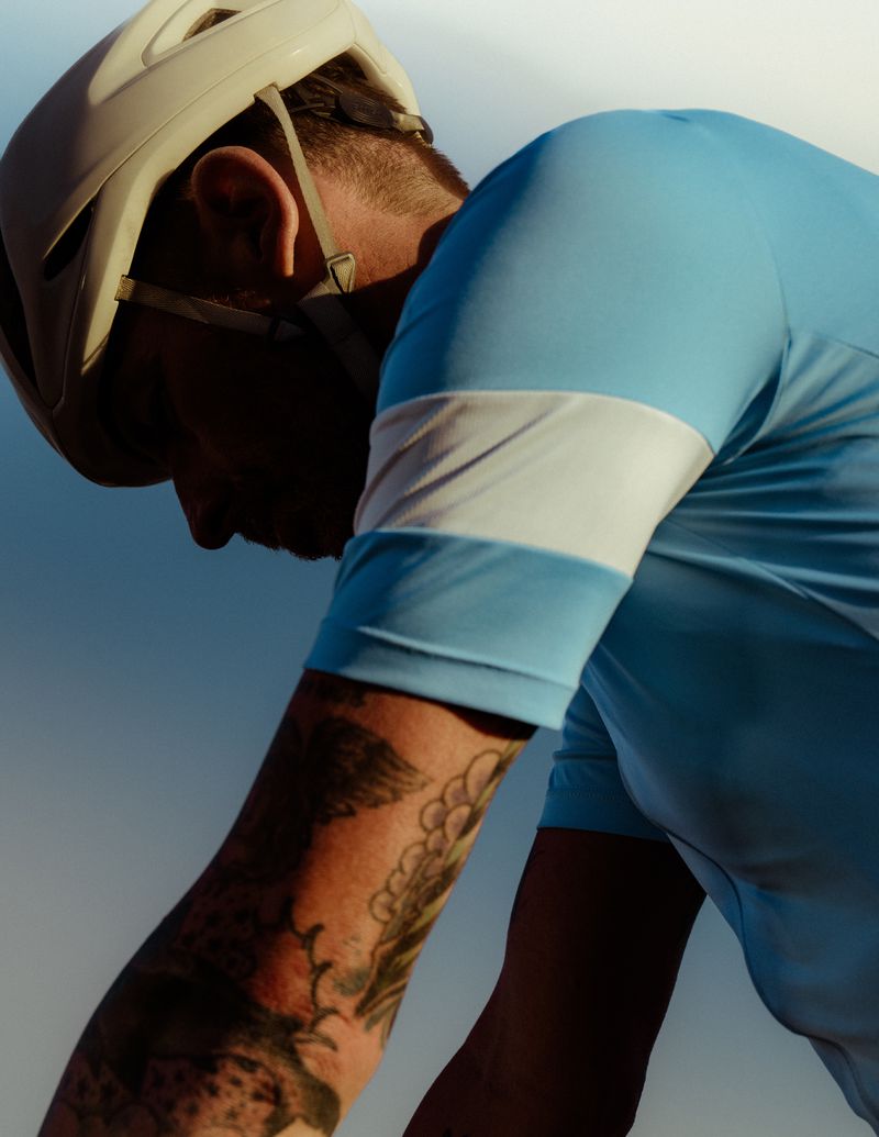 Cyclist wearing a helmet and blue cycling jersey in a close-up shot against a bright sky; arm tattoos are visible on the cyclist.