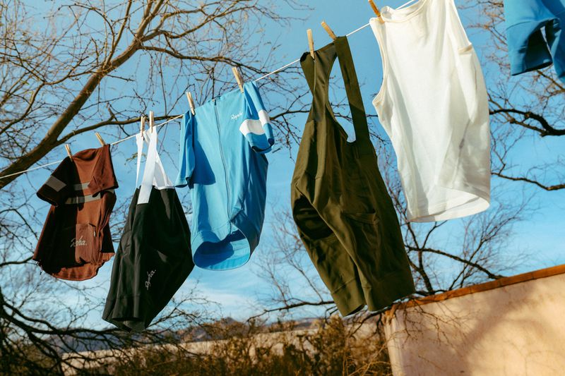 Cycling apparel hangs on a clothesline against a clear blue sky and bare tree branches, drying in the sunlight after washing.