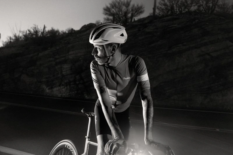 Cyclist wearing a white helmet and cycling jersey leaning over handlebars at night with a rocky background