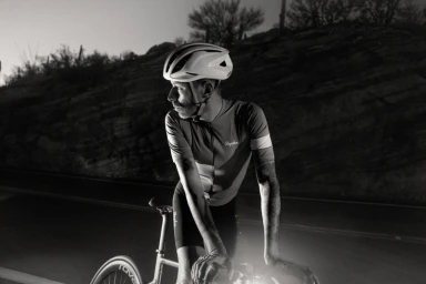 Cyclist wearing a white helmet and cycling jersey leaning over handlebars at night with a rocky background