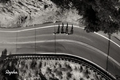 Overhead aerial view of four cyclists riding in a line along a winding mountain road, captured in high-contrast monochrome.