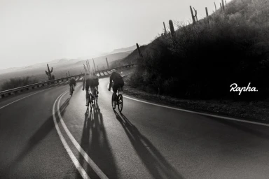 Monochrome image of four cyclists riding road bikes on a curving desert highway past saguaro cacti, photographed by Mark Mahaney.