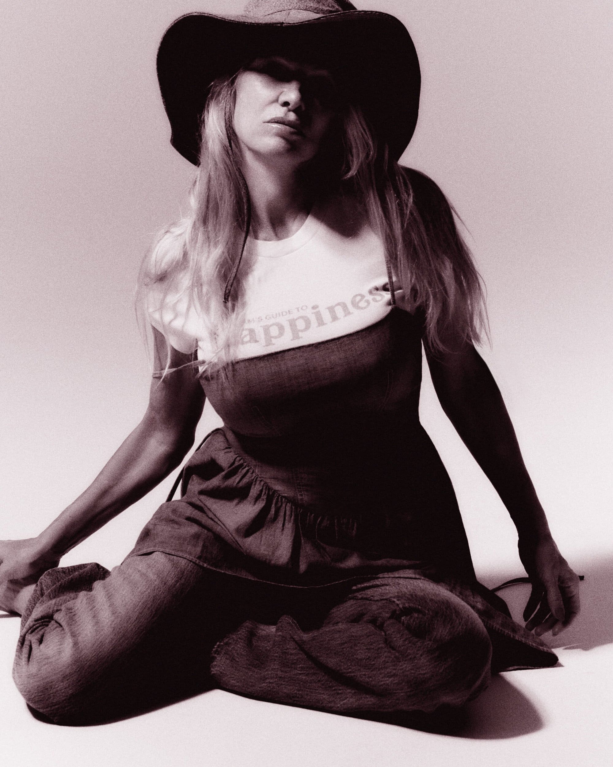 Blonde woman in a hat and overalls sitting cross-legged on the floor, wearing a white t-shirt that says, 'happiness'. Fashion.