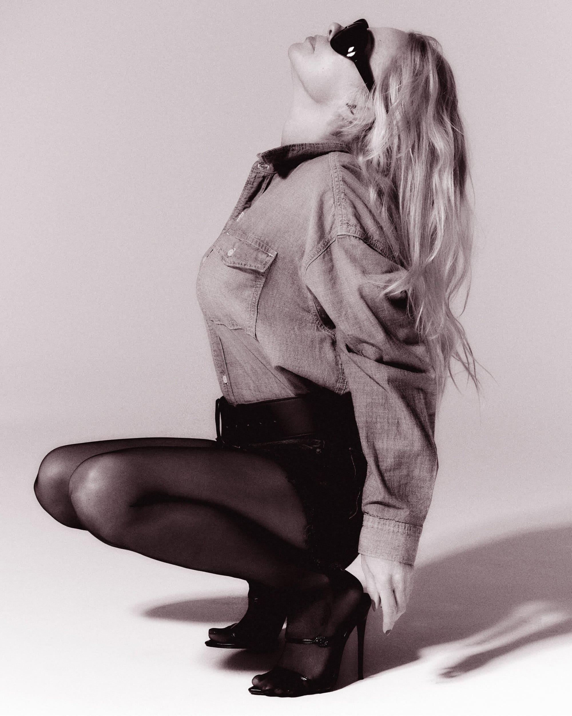 Model in denim shirt and dark stockings crouching against a neutral background wearing sunglasses.