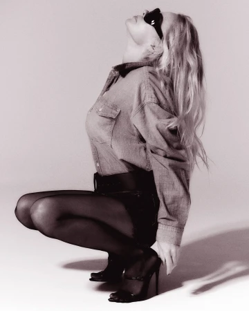 Model in denim shirt and dark stockings crouching against a neutral background wearing sunglasses.