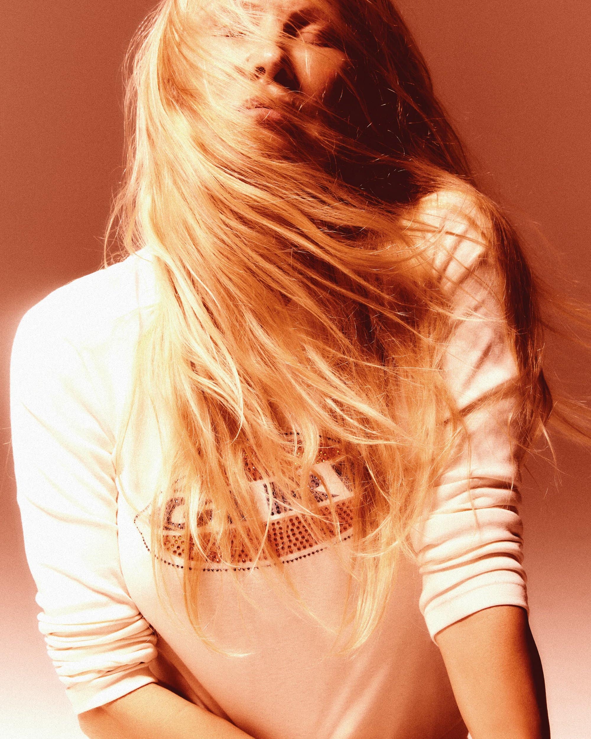 Portrait of a blonde woman with windblown hair, wearing a white top, eyes closed, against a plain, diffused background.