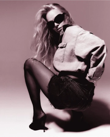 Model wearing dark sunglasses, denim shirt, and shorts crouching against a grey studio seamless backdrop for RE/DONE.