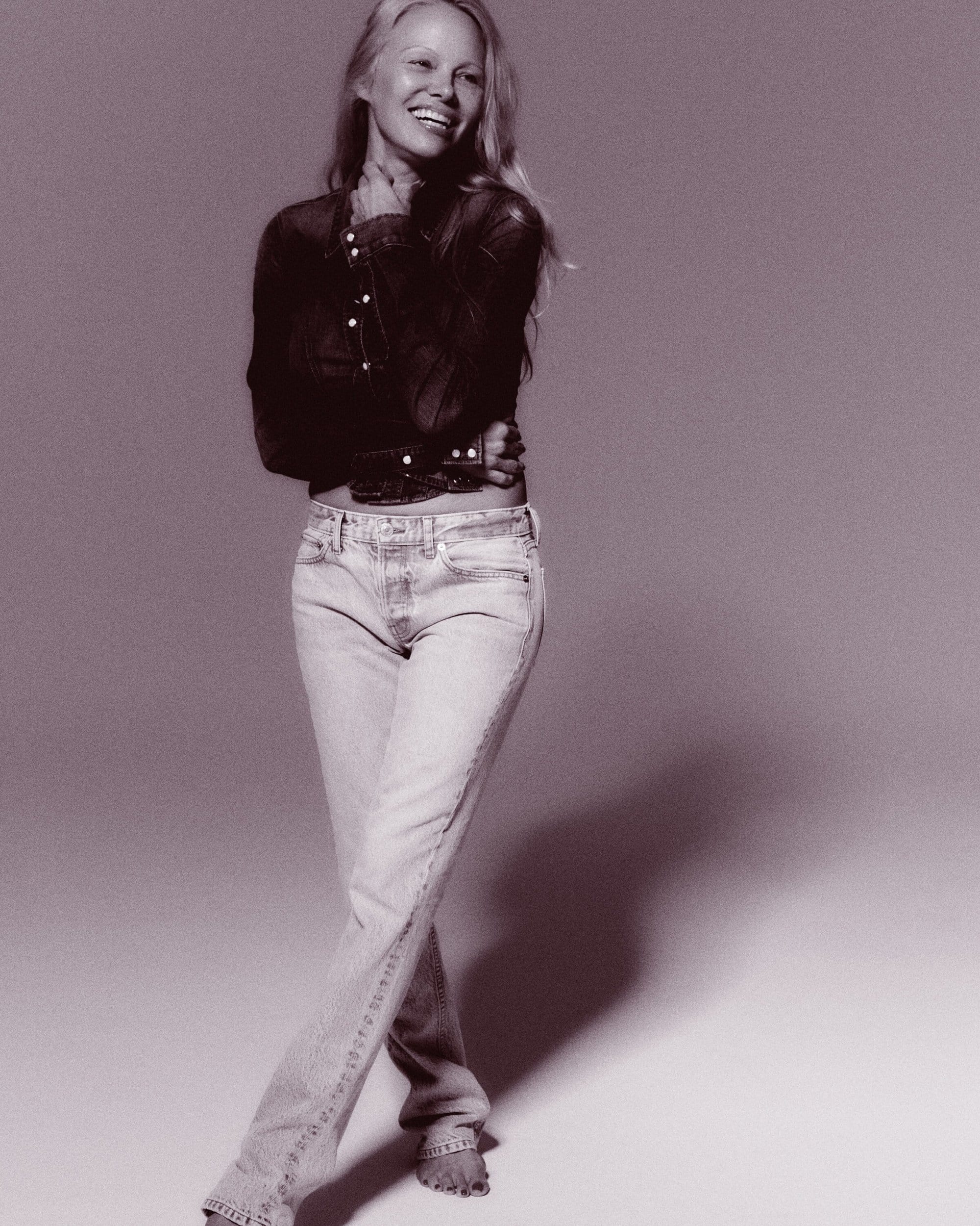 Pamela Anderson smiles while wearing a dark denim button-down shirt and light-wash straight-leg jeans in a studio portrait.