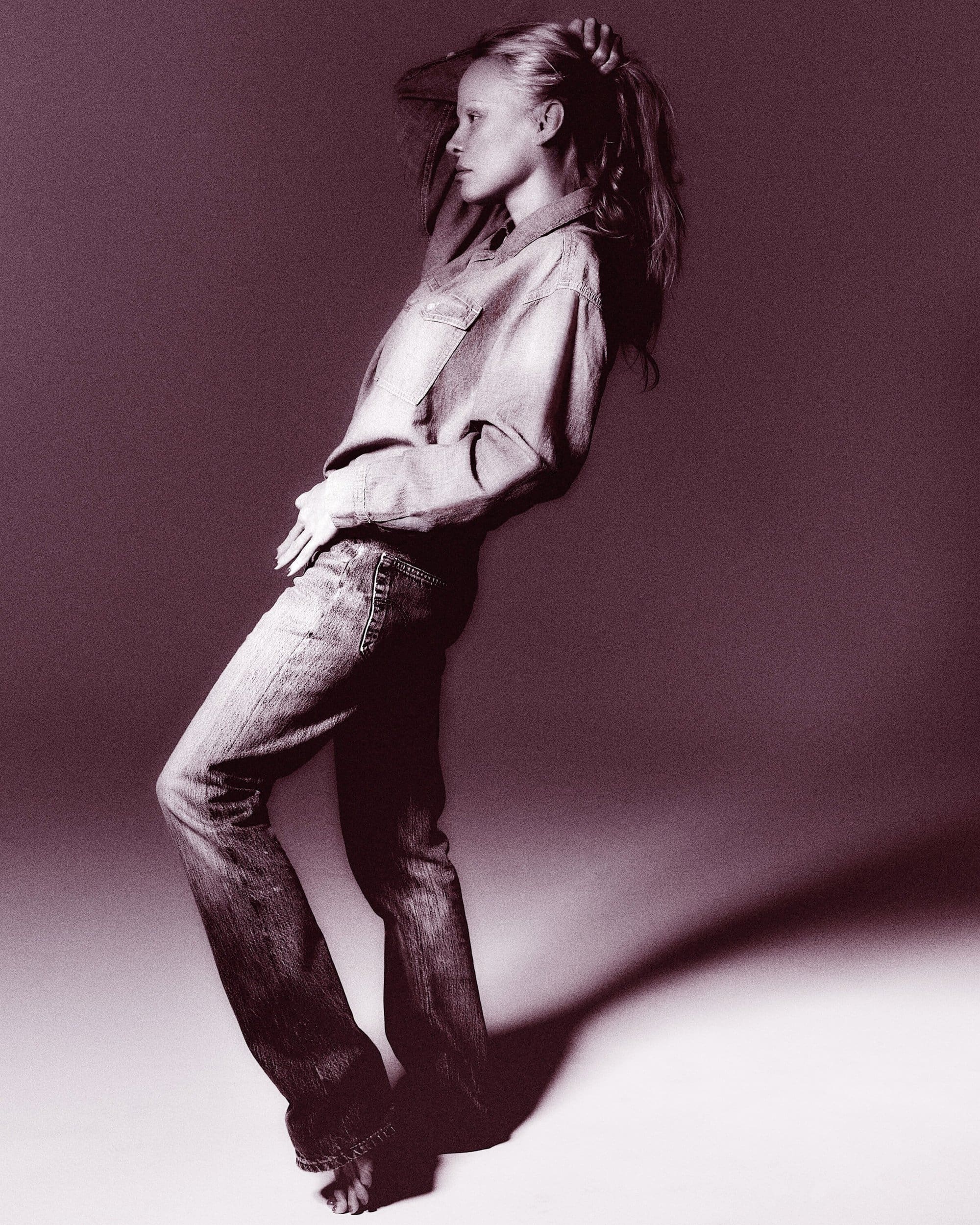 Blonde woman in denim shirt and jeans poses barefoot against a solid backdrop in a studio setting with soft lighting, fashion portrait.